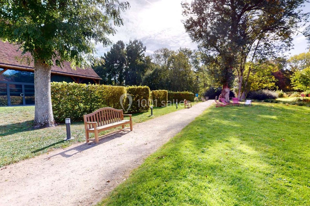 Allée bordée de bancs en bois, haies taillées et pelouse, menant à un bâtiment avec toit en pente sous un ciel dégagé.