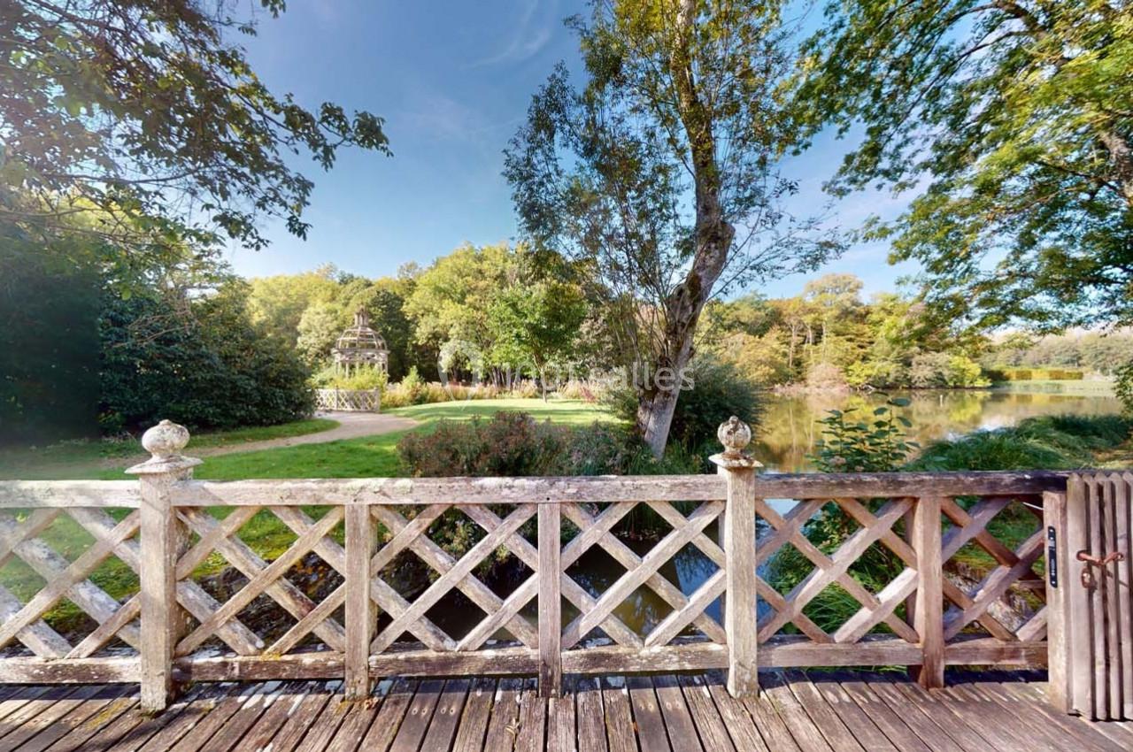 Vue depuis une terrasse en bois avec une balustrade, donnant sur un parc arboré et un étang sous un ciel dégagé.