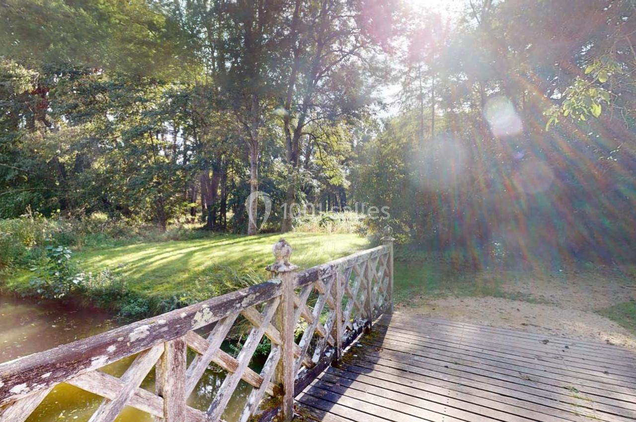Pont en bois avec balustrades décoratives traversant un petit cours d'eau, entouré de verdure et baigné de lumière.