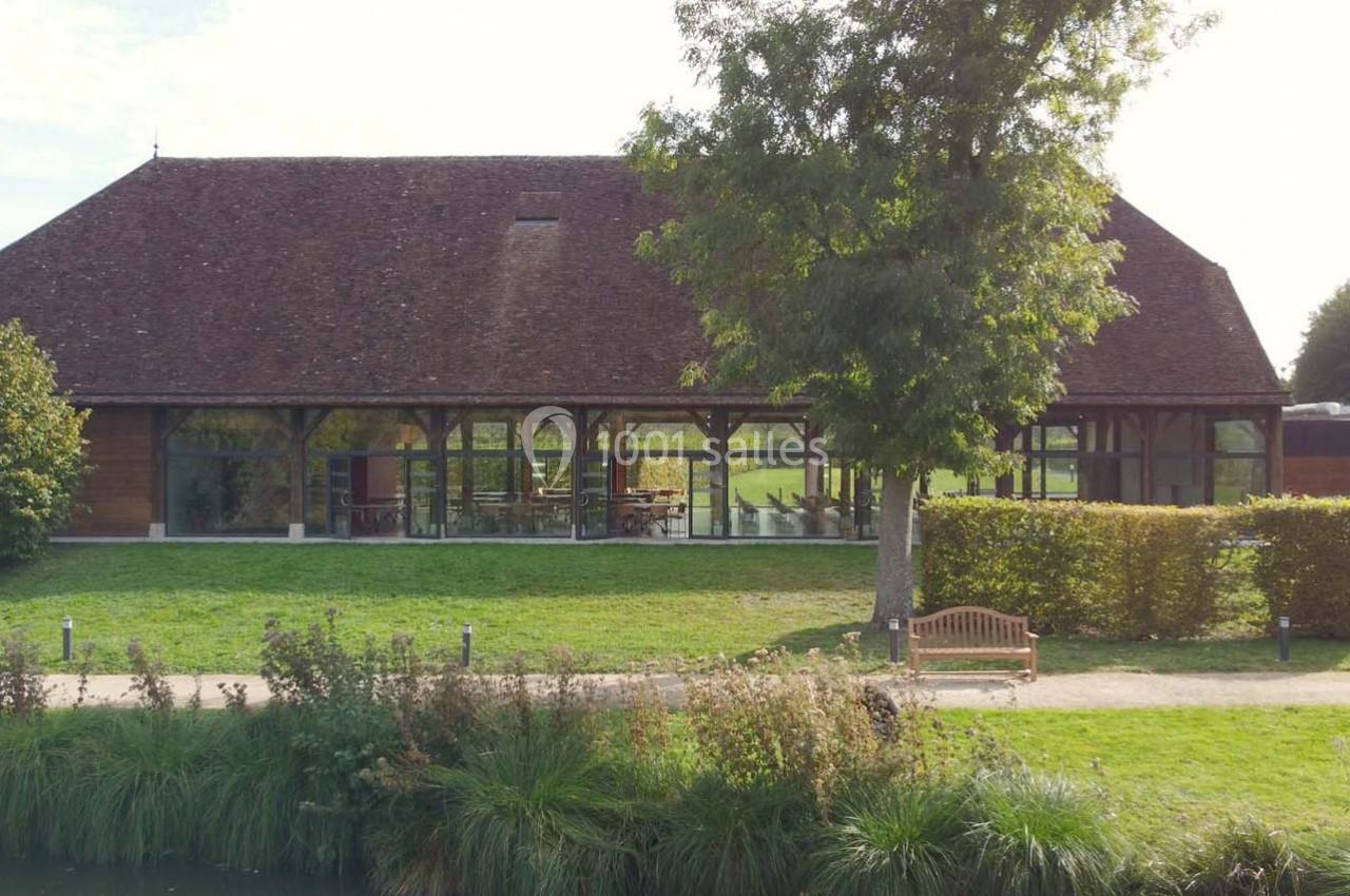 Bâtiment en bois avec de grandes baies vitrées, entouré de verdure et d'un chemin longeant un étang.