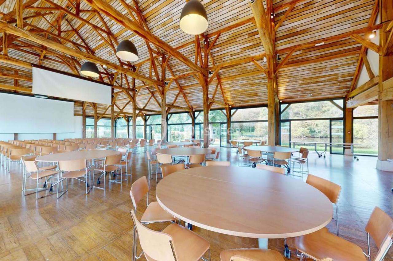 Salle lumineuse avec charpente en bois, tables rondes et chaises, équipée d'un écran de projection.