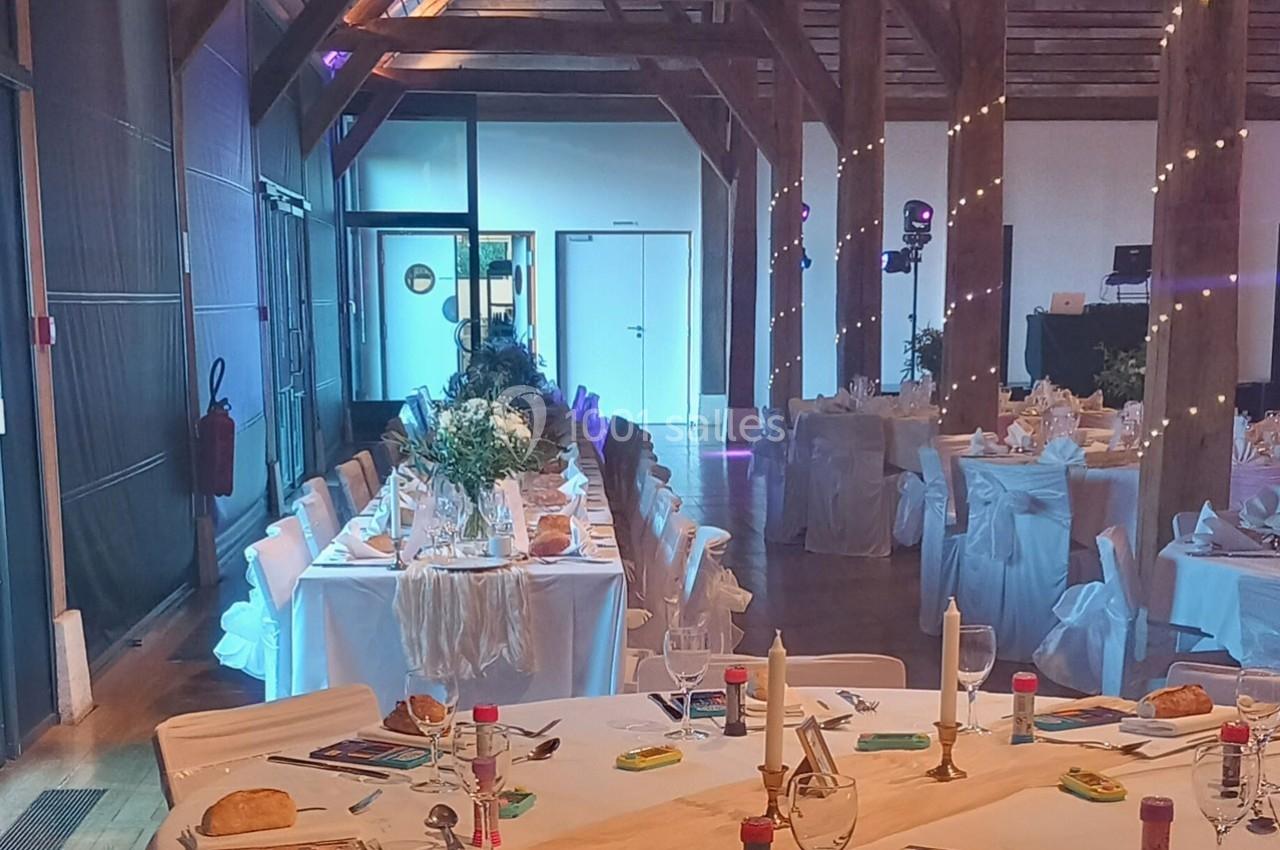 Salle de réception décorée pour un événement, avec tables dressées, chaises blanches et guirlandes lumineuses suspendues.