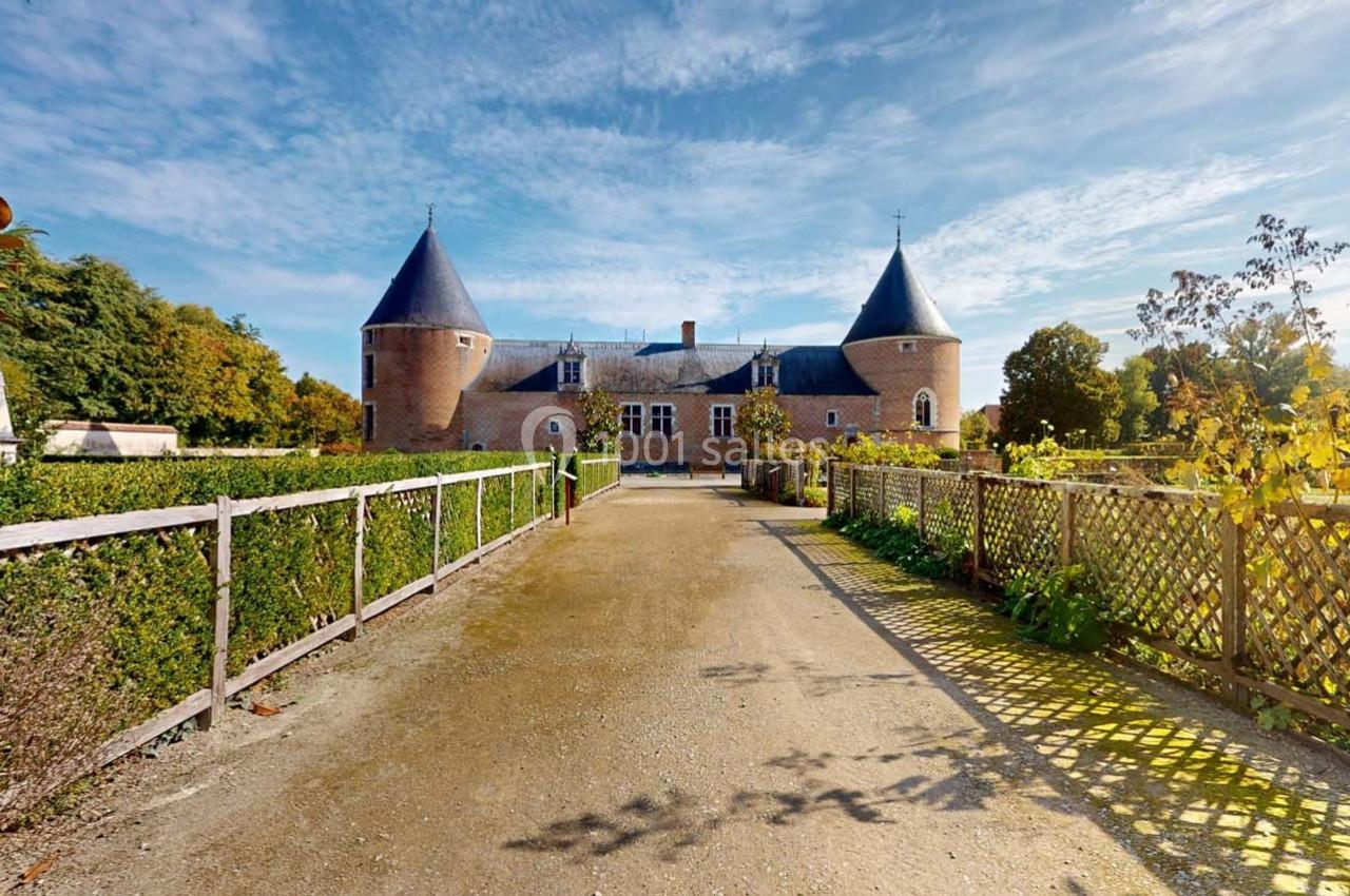 Allée bordée de haies menant à un château en briques rouges avec deux tours, sous un ciel dégagé.