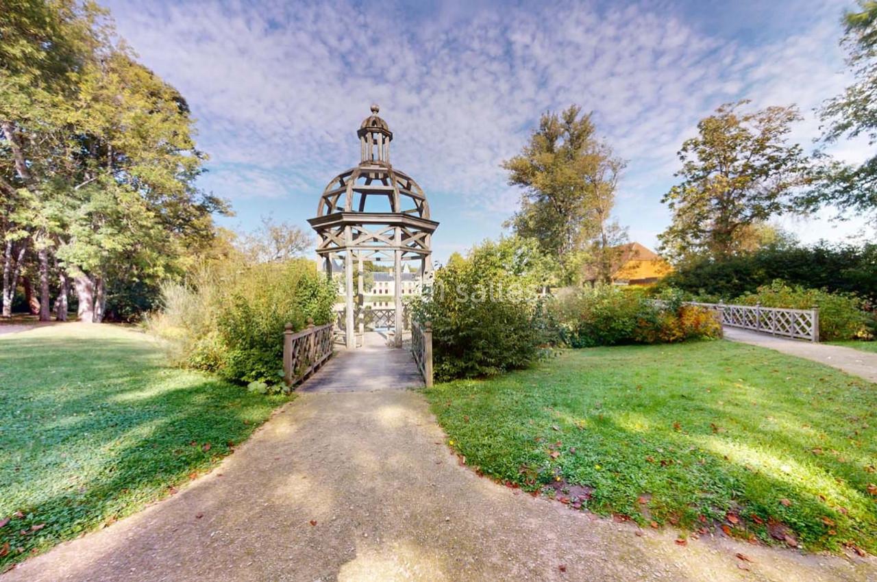 Kiosque en bois au centre d'un jardin verdoyant, entouré de chemins et de végétation sous un ciel partiellement nuageux.
