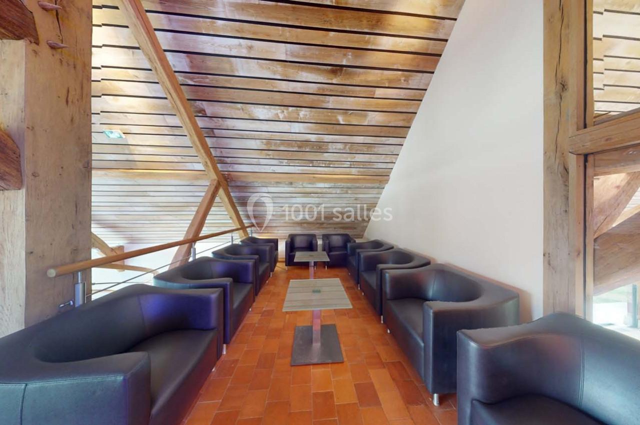 Espace lounge avec fauteuils noirs disposés autour de petites tables sur un sol en carreaux rouges, sous un plafond en bois.