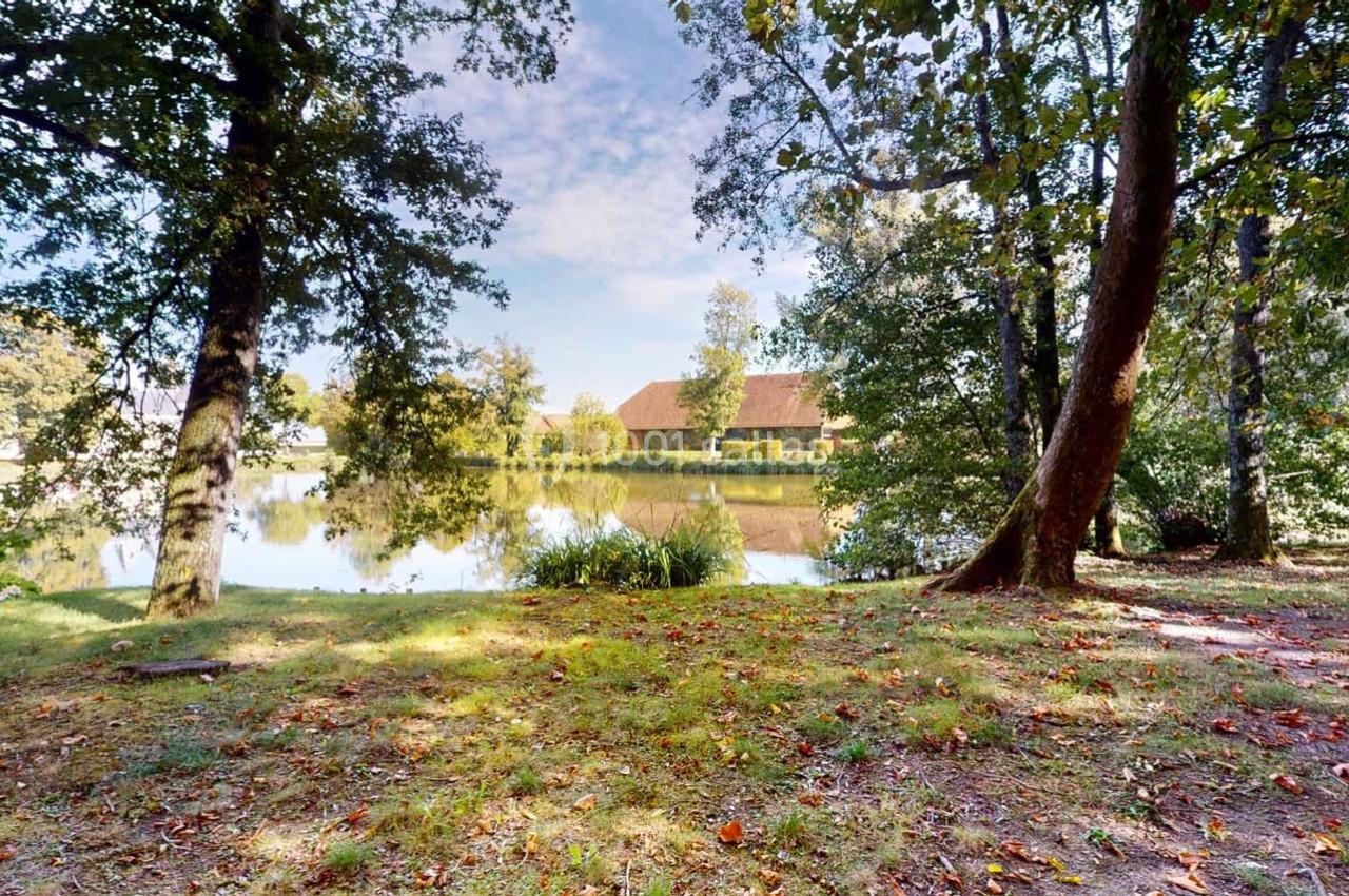 Vue d'un étang entouré d'arbres avec une maison en arrière-plan sous un ciel partiellement nuageux.