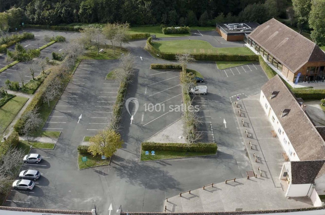 Vue aérienne d'un parking partiellement occupé, entouré de végétation et de bâtiments à toit en tuiles.