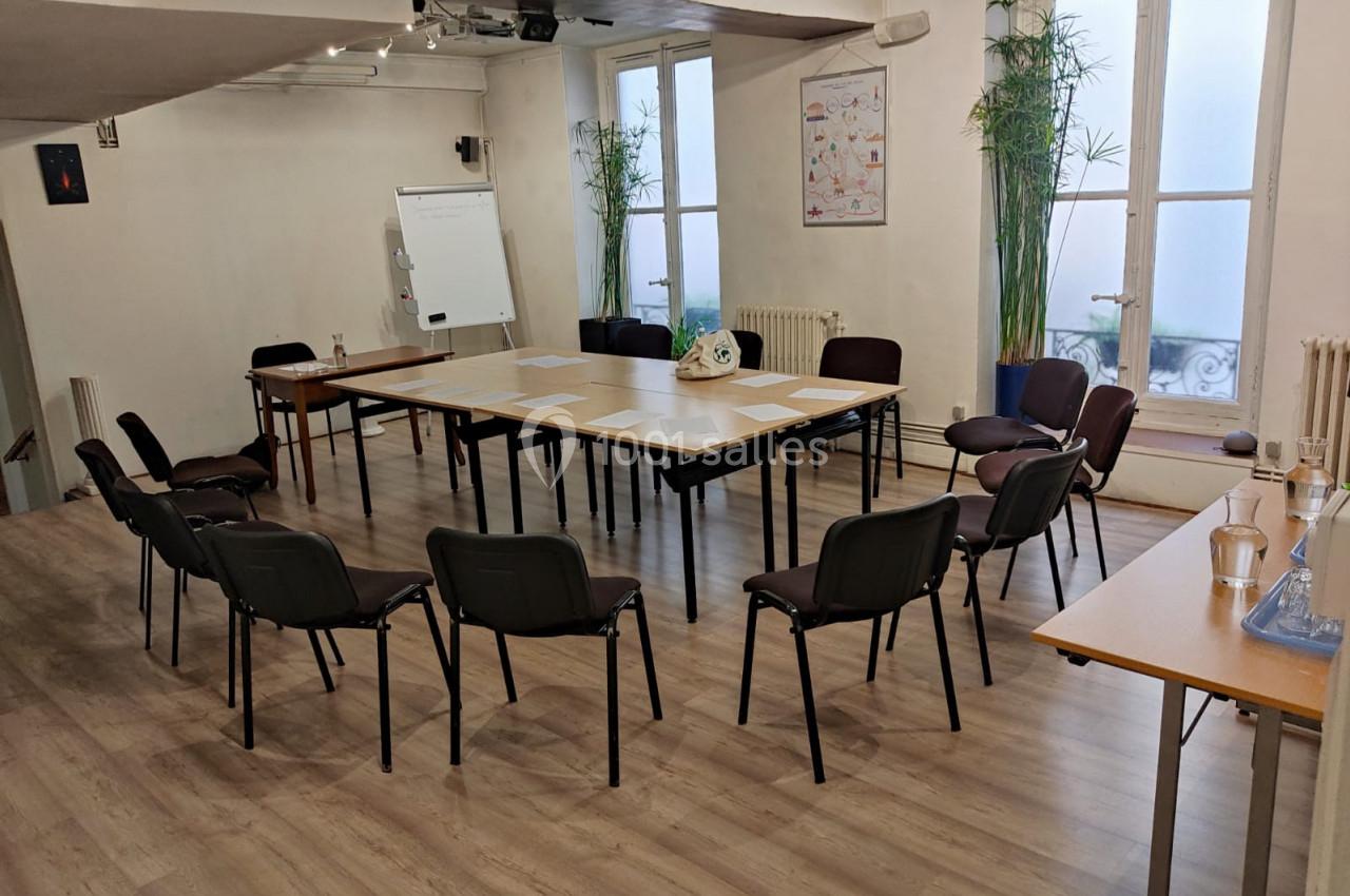 Salle de réunion lumineuse avec tables disposées en U, chaises autour, tableau blanc et plantes en arrière-plan.