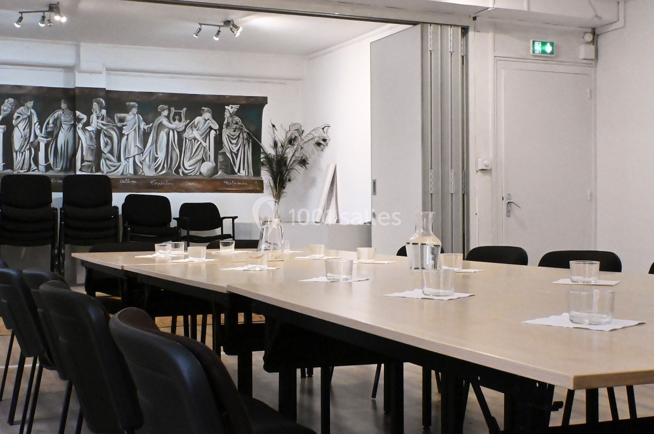 Salle de réunion avec une grande table, des chaises noires, des carafes d'eau et un tableau représentant des figures…