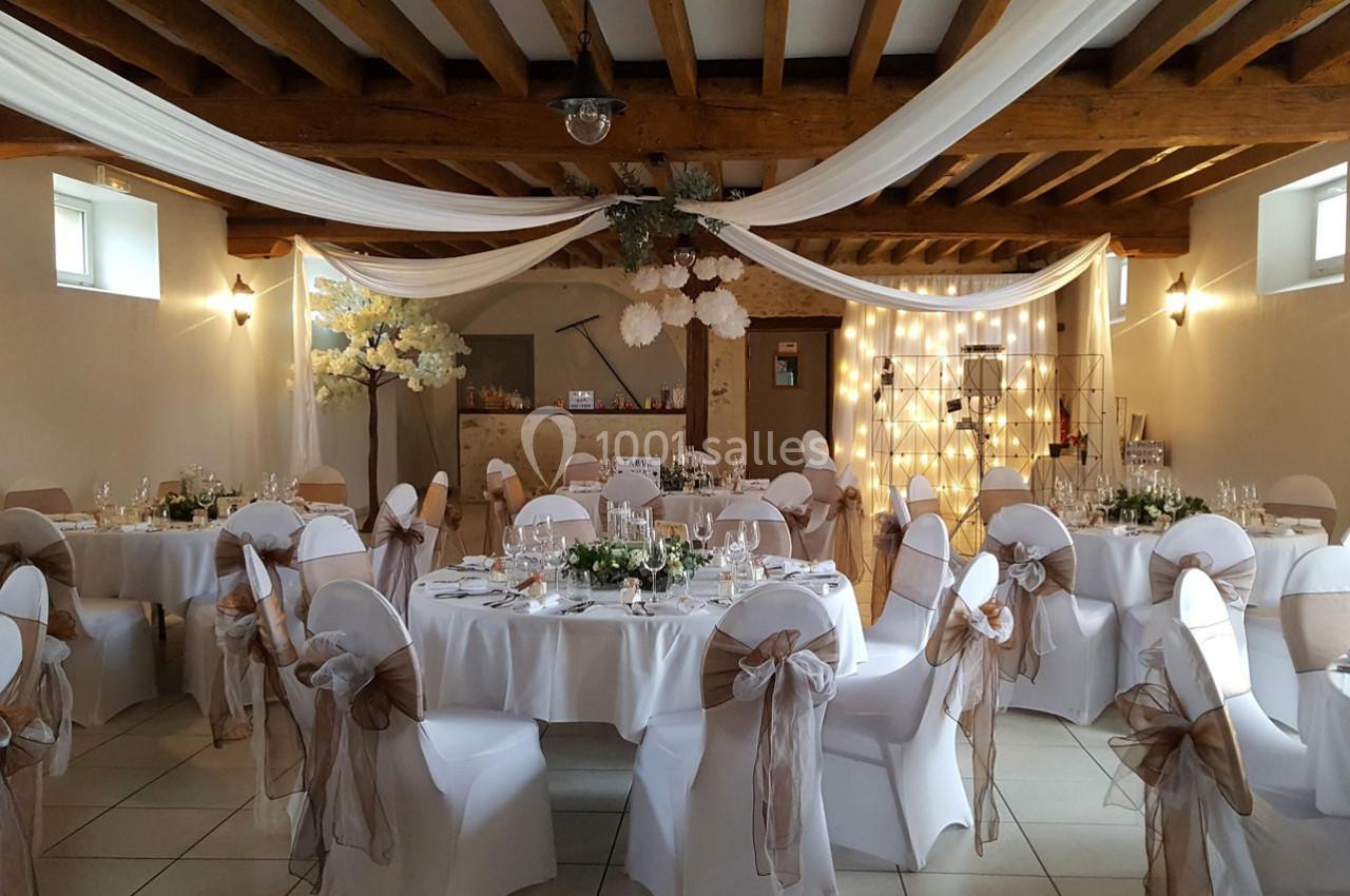 Salle de réception décorée pour un mariage, avec tables rondes, chaises ornées de nœuds et drapés suspendus au plafond.
