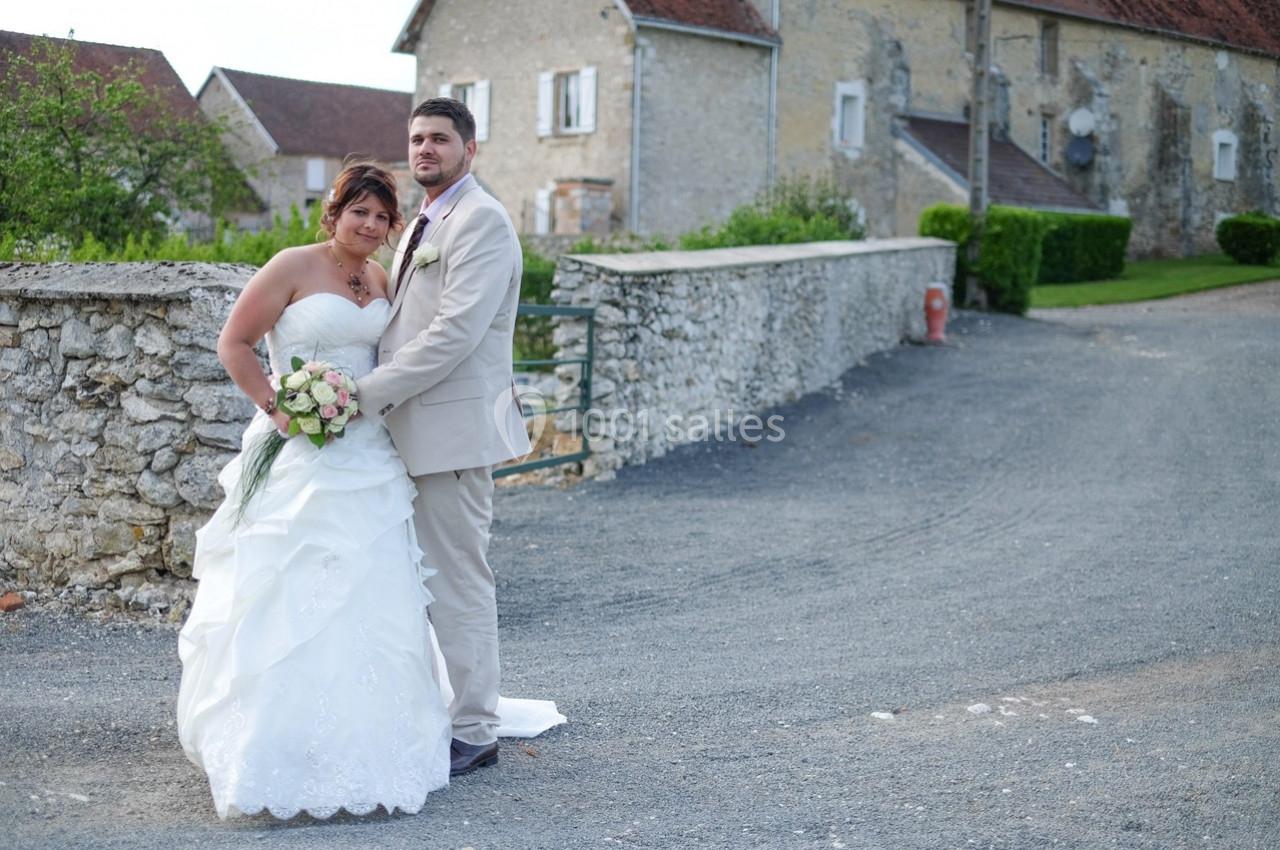 Un couple en tenue de mariage pose devant un mur de pierre, avec des bâtiments en arrière-plan.