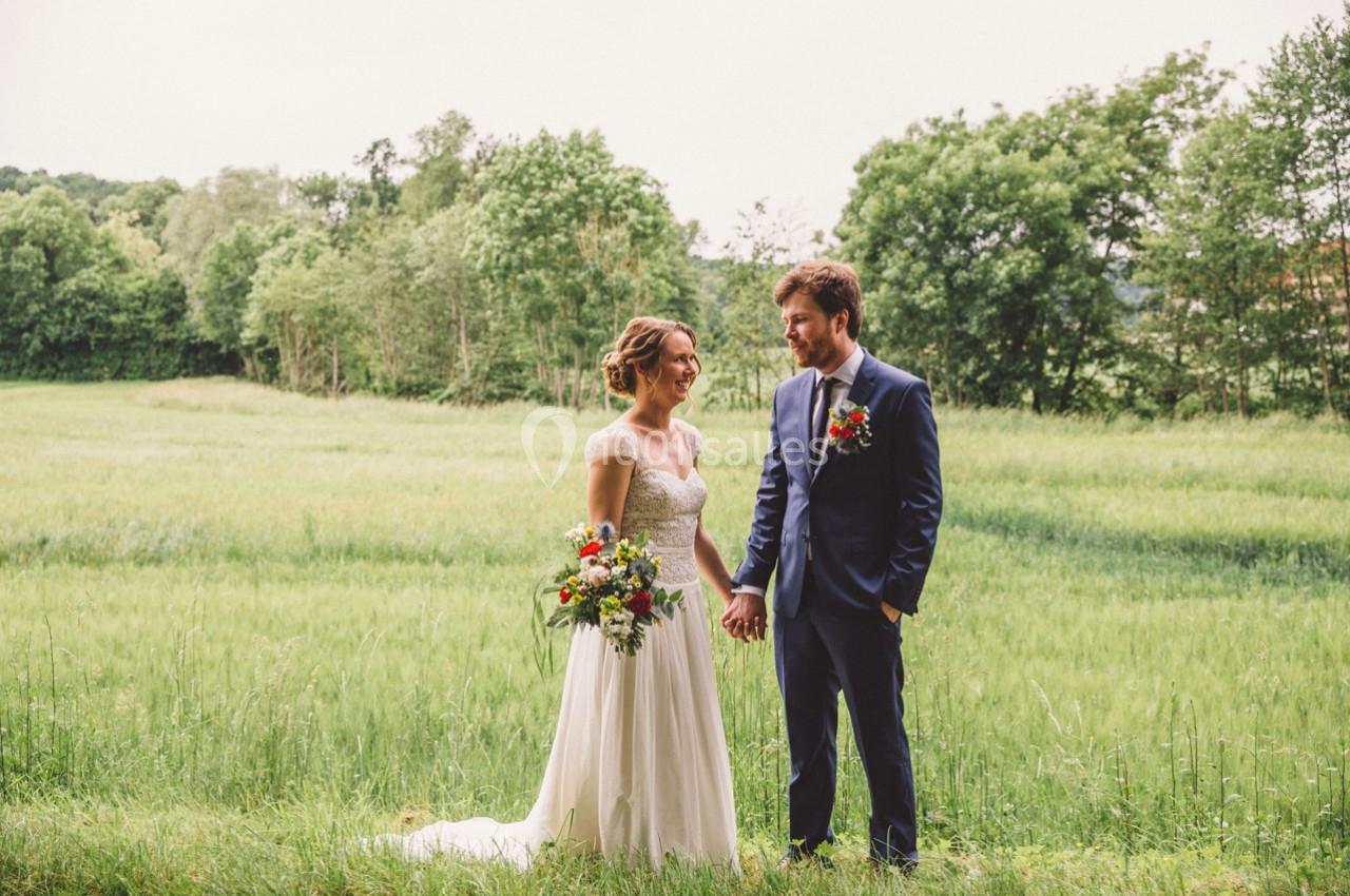 Un couple en tenue de mariage se tient la main dans un champ verdoyant bordé d'arbres.