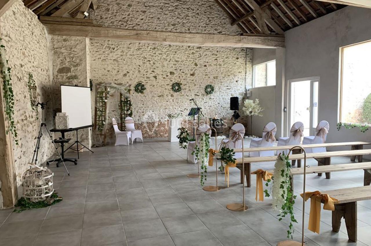 Salle de réception rustique décorée pour un événement avec chaises blanches, fleurs et pupitre, dans un cadre en pierre.