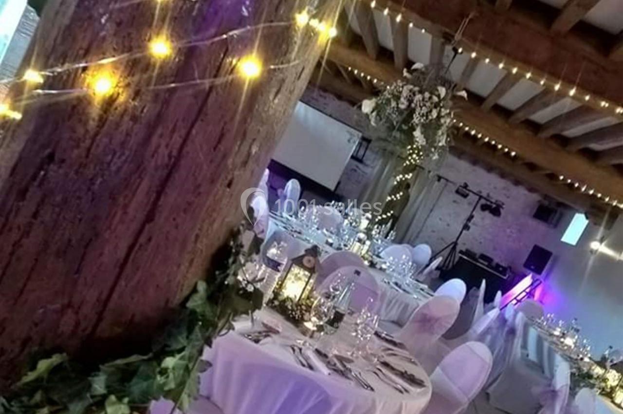 Salle de réception décorée avec des guirlandes lumineuses, des tables rondes dressées et des chaises recouvertes de housses…