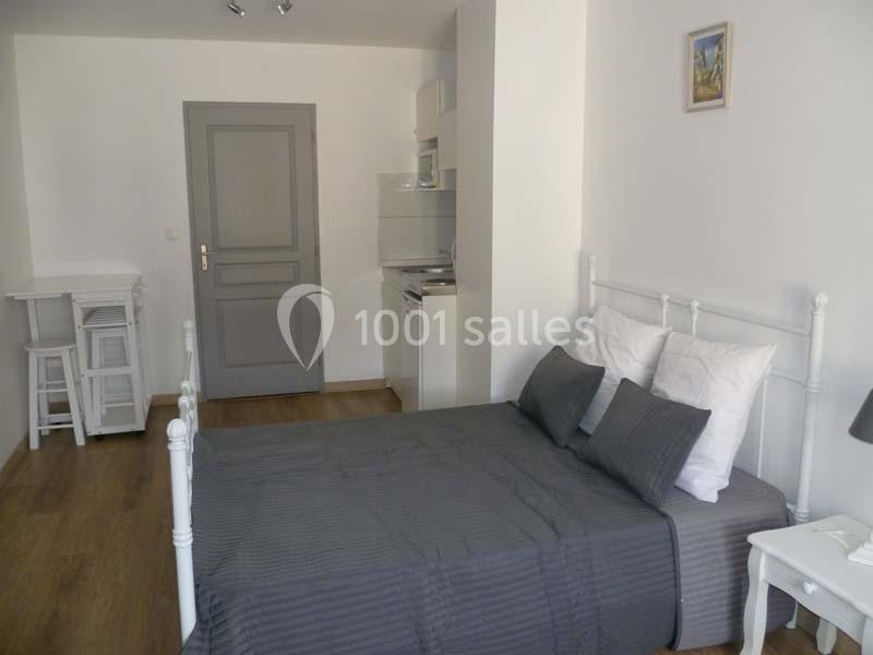 Chambre lumineuse avec lit double, linge de lit gris, kitchenette équipée et mobilier blanc dans un espace moderne.