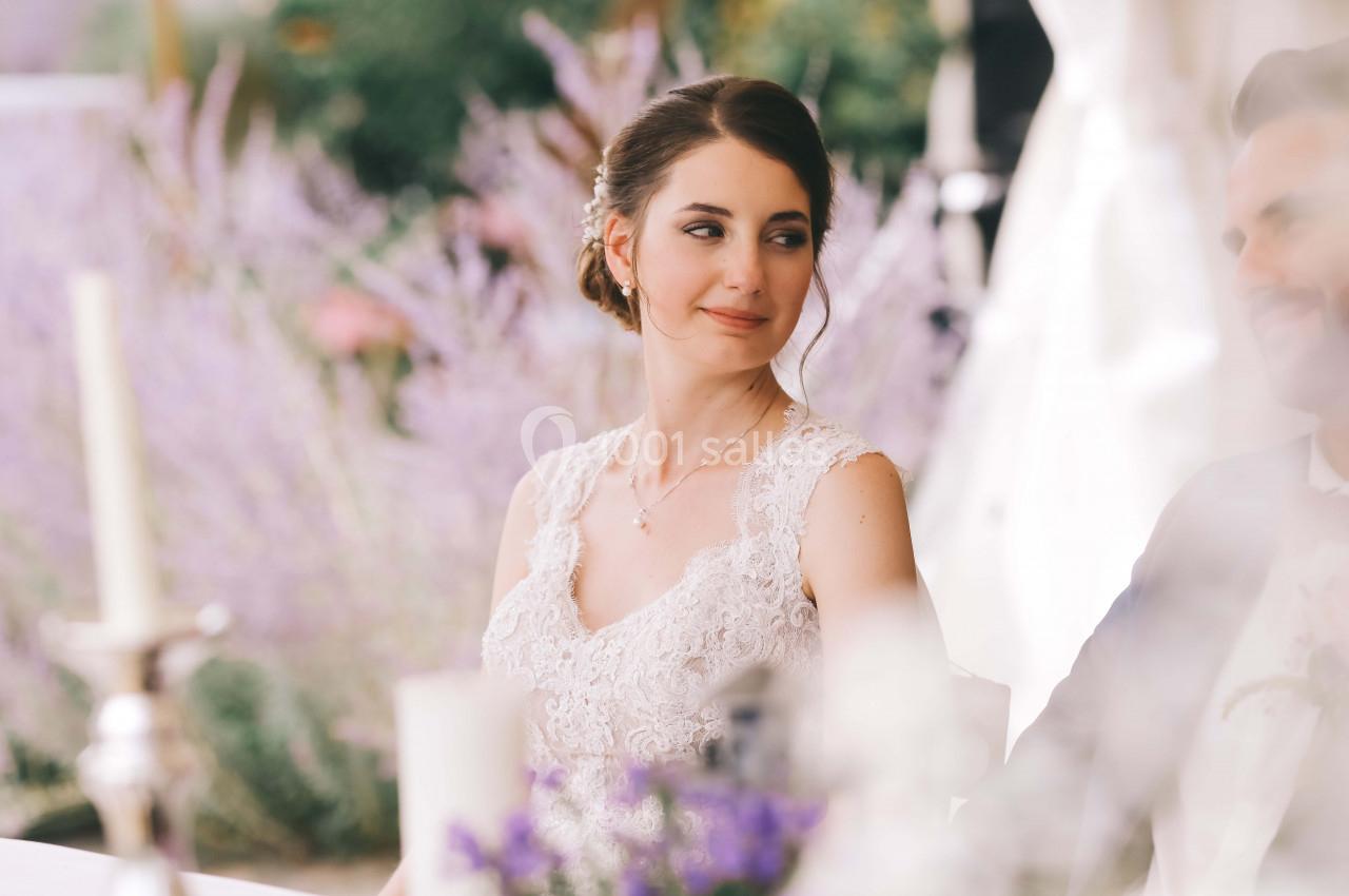 Une femme en robe de mariée assise à une table, regardant sur le côté, avec un fond de lavande flou.