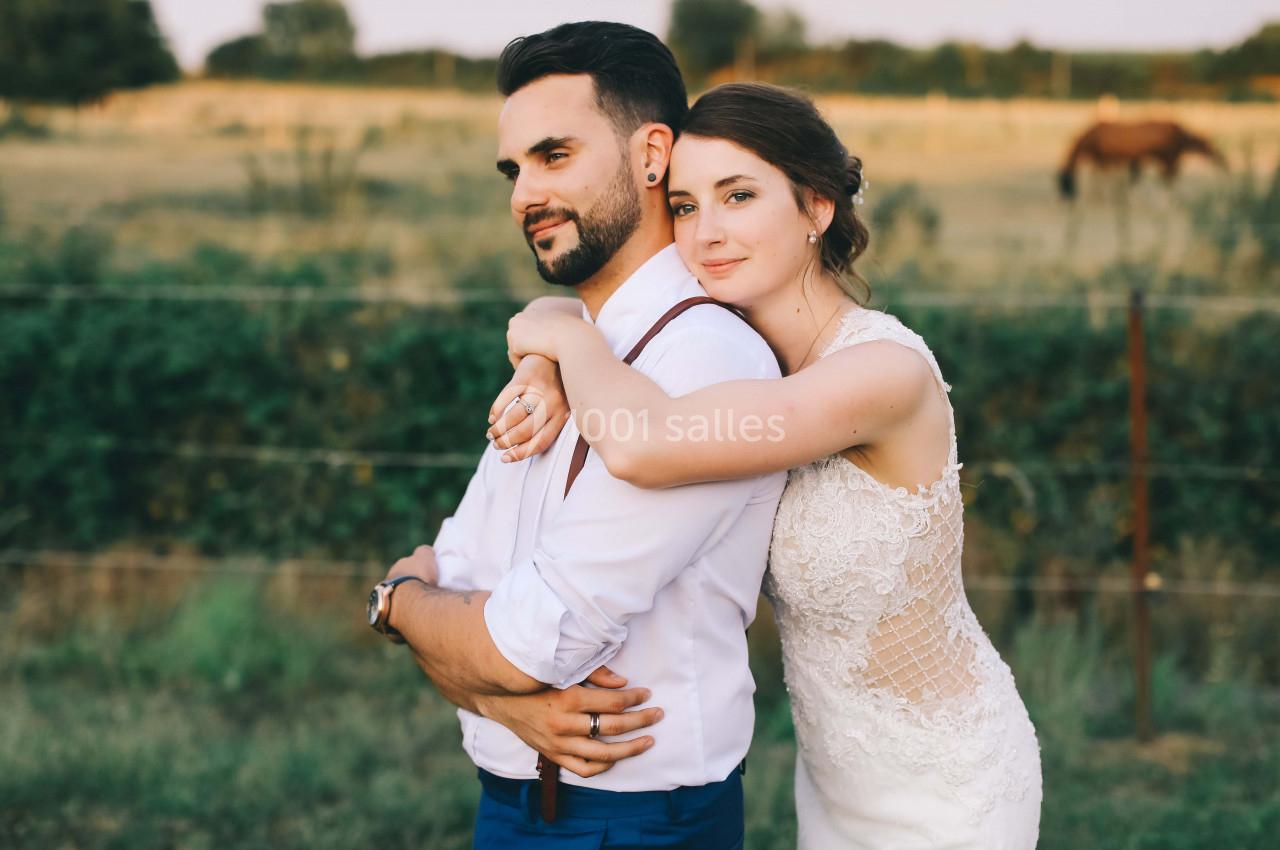 Un couple en tenue de mariage pose dans un champ, la mariée entourant le marié de ses bras.