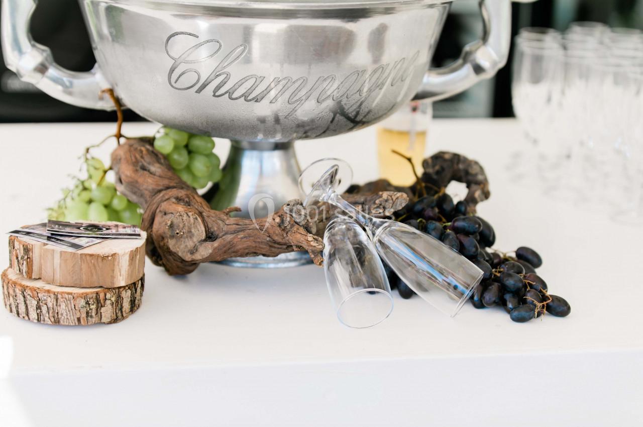 Seau à champagne en métal entouré de raisins, verres renversés et décorations en bois sur une table blanche.