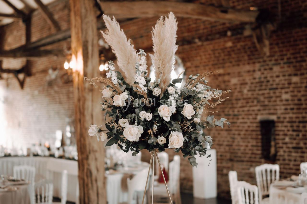 Centre de table floral avec pampas et roses blanches dans une salle de réception en briques avec poutres apparentes.