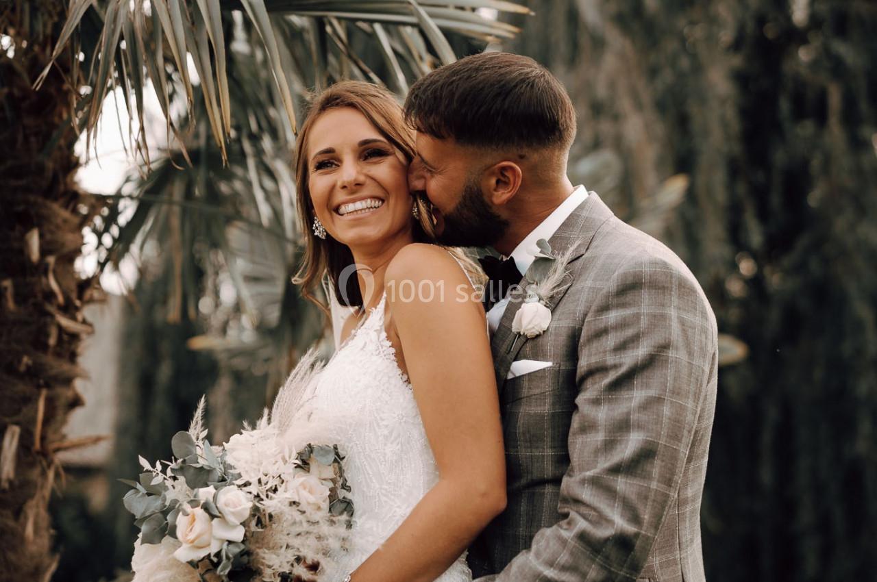 Un couple de mariés souriant, l'homme en costume gris embrasse la femme en robe blanche tenant un bouquet, devant des arbres.