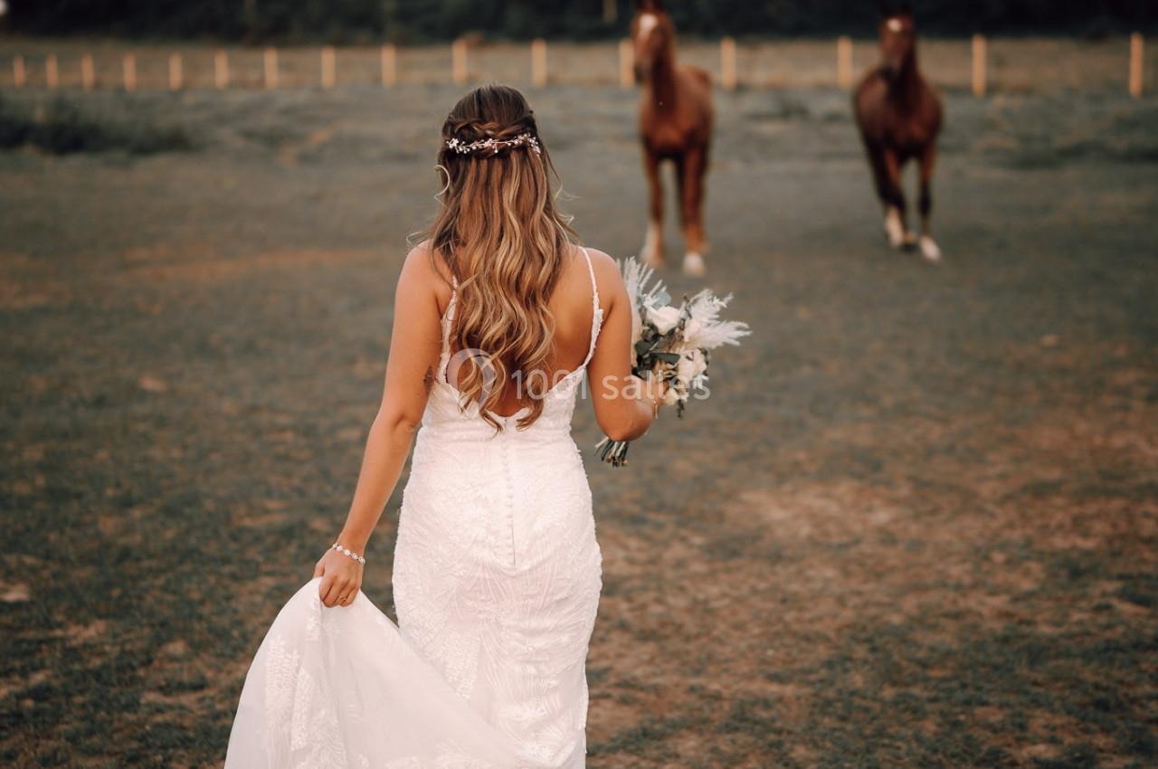 Une femme en robe de mariée marche dans un champ, tenant sa traîne et un bouquet, avec deux chevaux en arrière-plan.