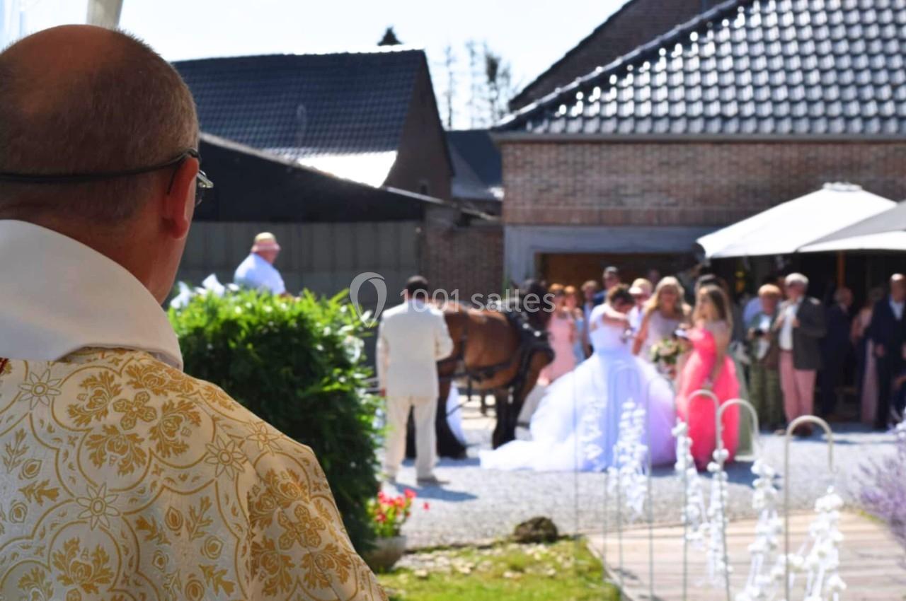 Un prêtre observe une cérémonie de mariage en extérieur, avec des invités rassemblés près d'une calèche.