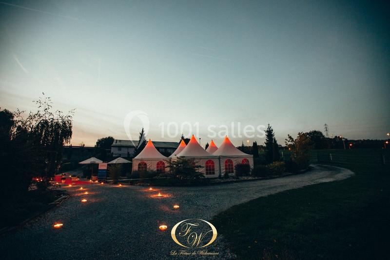 Tentes blanches éclairées au crépuscule, entourées de verdure et d'un chemin gravillonné illuminé.