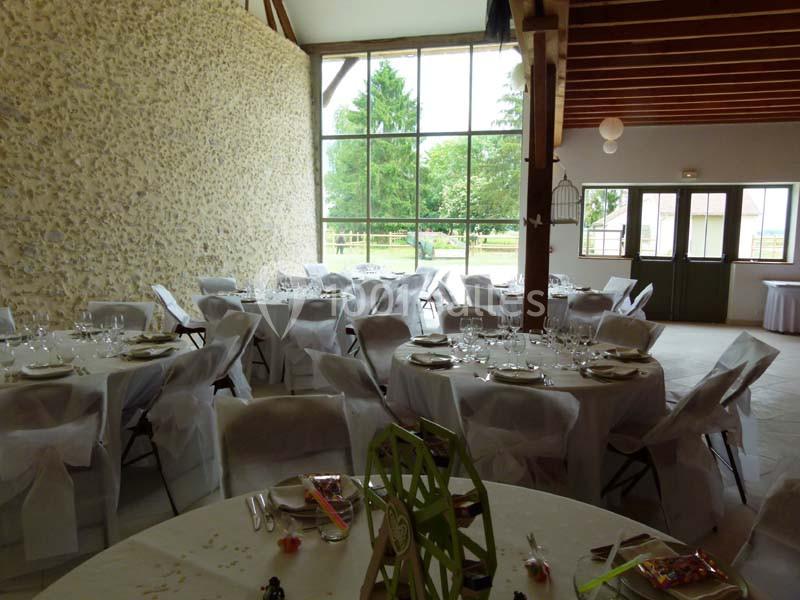 Salle de réception lumineuse avec tables rondes dressées, nappes blanches et grandes baies vitrées donnant sur un jardin.