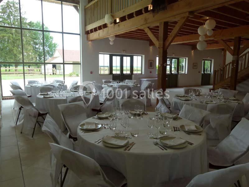 Salle de réception lumineuse avec tables rondes dressées, nappes blanches et vue sur l'extérieur par de grandes baies…