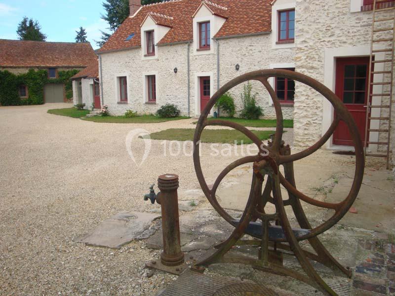 Vieille pompe à eau en métal devant une maison en pierre avec toit en tuiles rouges et cour en gravier.