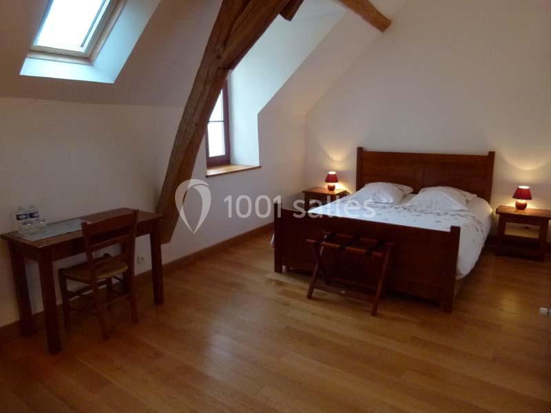 Chambre mansardée avec lit double en bois, bureau, chaise, parquet et lumière naturelle provenant d'une fenêtre de toit.