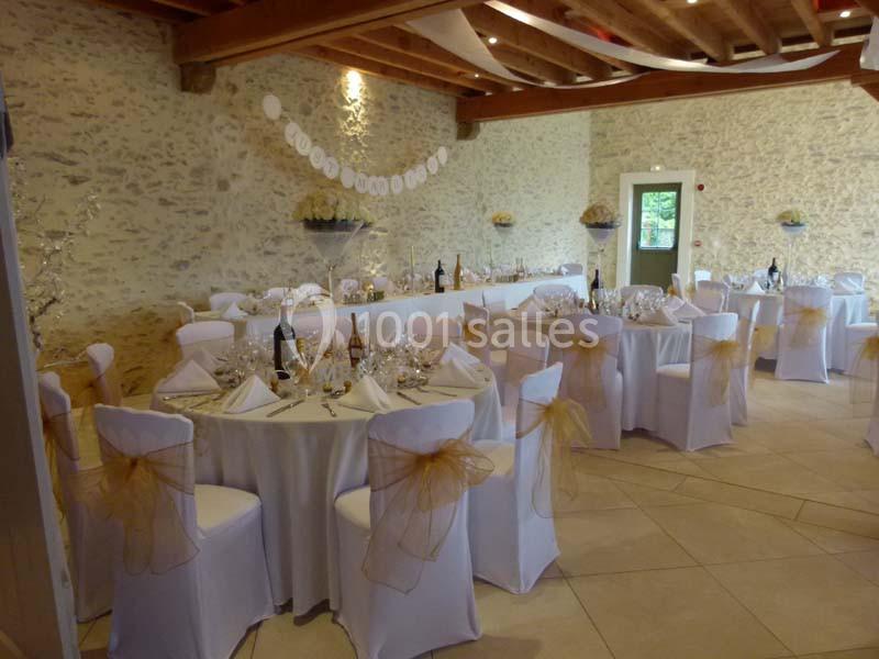 Salle de réception décorée pour un événement, avec des tables dressées, des chaises ornées de nœuds dorés et des murs en…