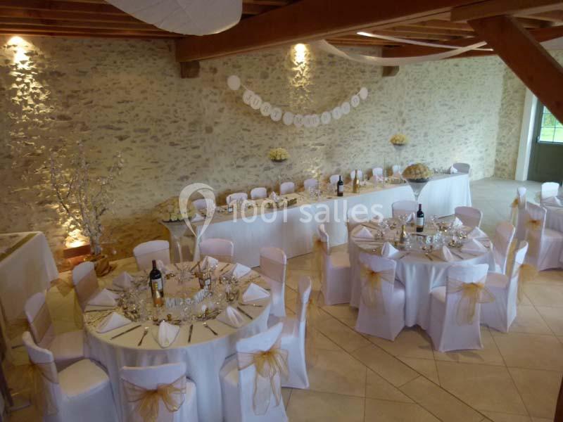 Salle de réception décorée avec des tables rondes et une table d'honneur, nappes blanches et chaises ornées de rubans dorés.