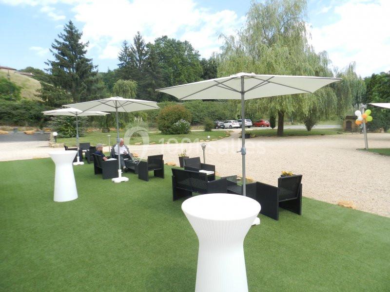 Terrasse extérieure avec tables, chaises et parasols blancs sur une pelouse synthétique, entourée de verdure.