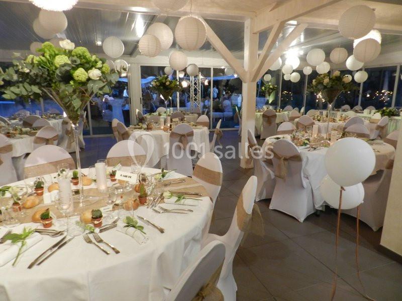 Salle de réception décorée avec des tables rondes, nappes blanches, chaises couvertes et arrangements floraux élégants.
