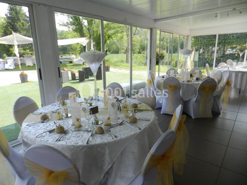 Salle de réception lumineuse avec tables rondes dressées, nappes blanches et chaises décorées de nœuds jaunes.