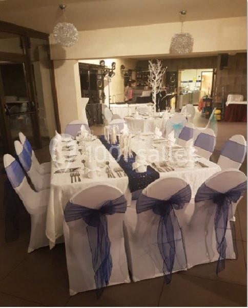 Table dressée pour un événement, avec nappes blanches, chaises décorées de nœuds bleus et centre de table élégant.