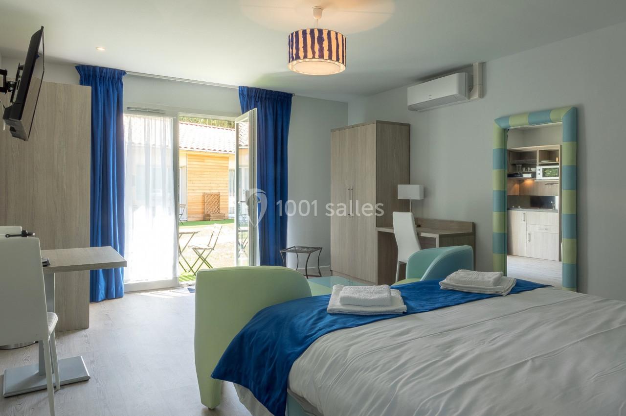 Chambre lumineuse avec lit double, bureau, kitchenette et accès à une terrasse extérieure par une porte vitrée.