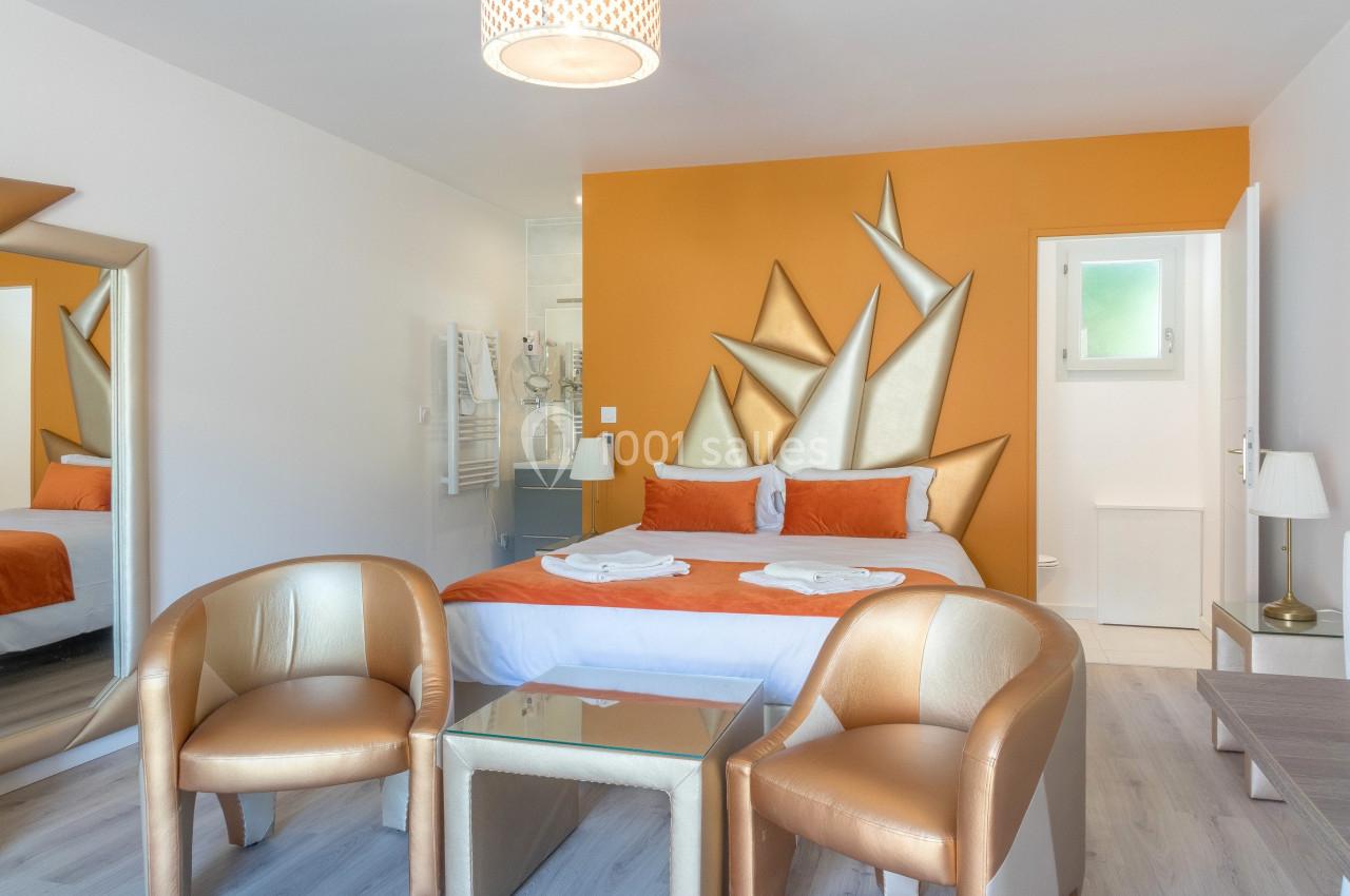 Chambre moderne avec lit double, mur orange décoratif, deux fauteuils dorés et lumière naturelle.