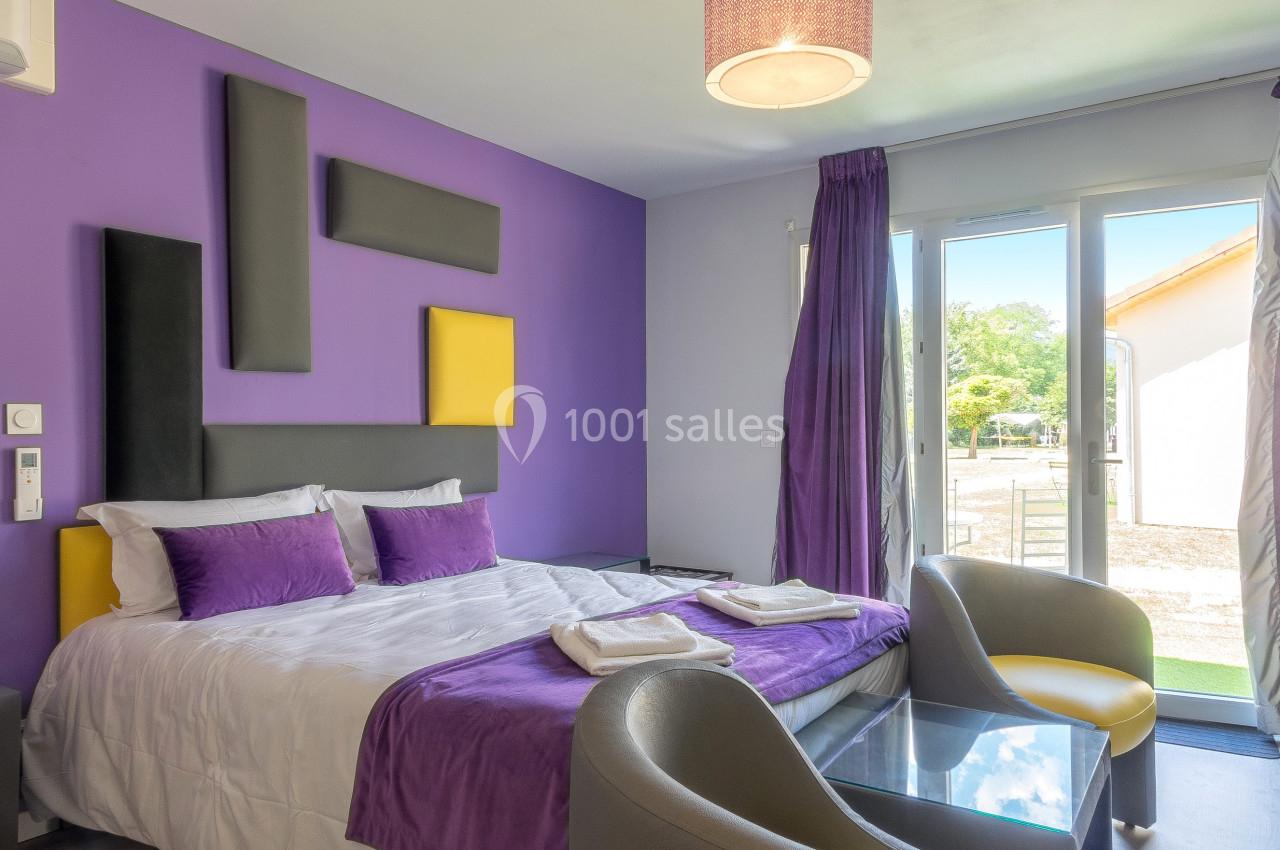 Chambre lumineuse avec lit double, murs violets, fauteuils gris, table basse en verre et grande baie vitrée donnant sur l…