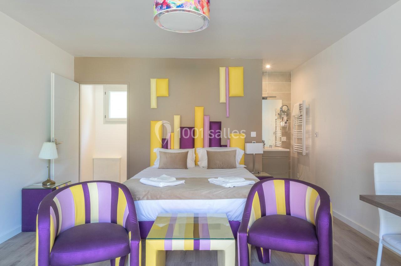 Chambre lumineuse avec lit double, chaises colorées, table basse et accès à une salle de bain moderne.