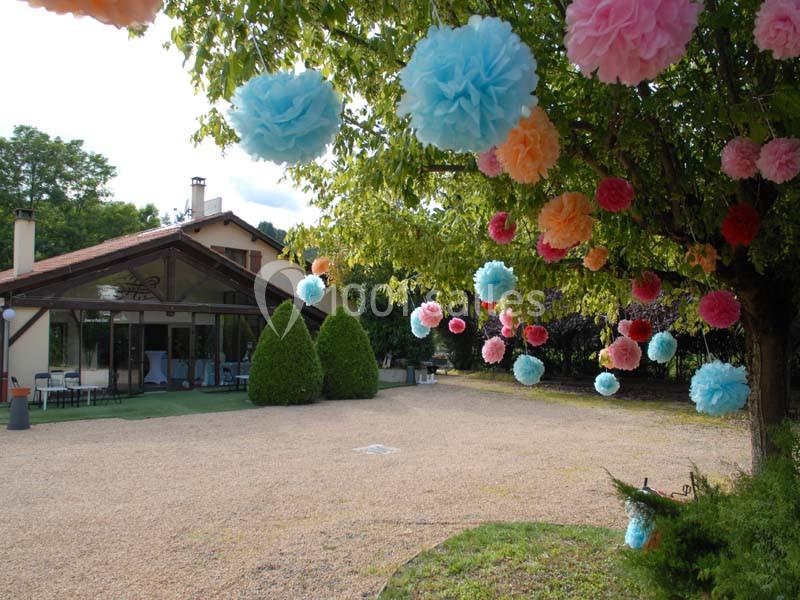 Pompons colorés suspendus à un arbre dans un jardin près d'une maison avec terrasse et pelouse.
