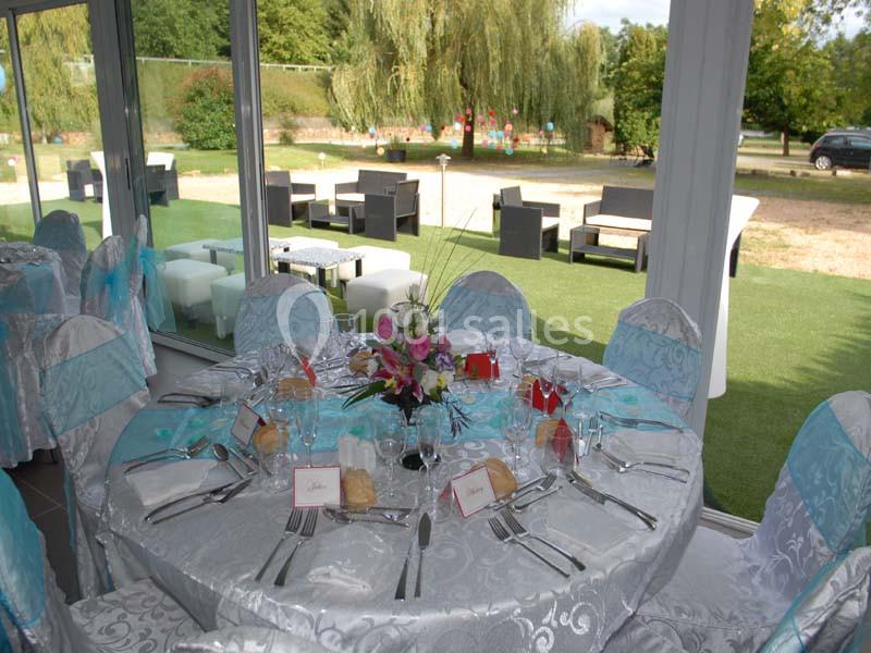 Table dressée avec nappes blanches, couverts, verres et centre de table fleuri, dans une salle lumineuse avec vue sur jardin.