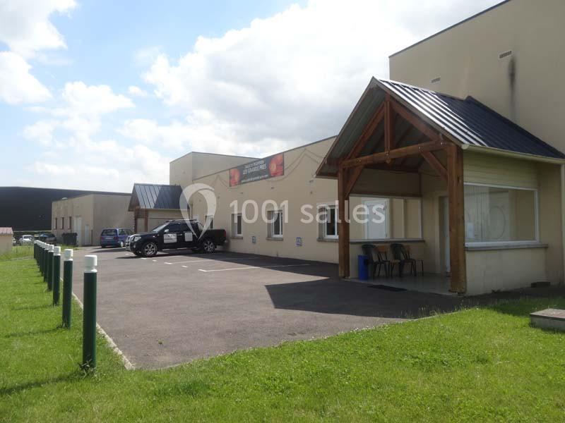 Bâtiment beige avec toit en tôle, parking asphalté et pelouse verte au premier plan, sous un ciel partiellement nuageux.