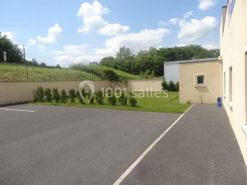 Parking asphalté avec un espace vert bordé d'arbustes, adjacent à un bâtiment beige sous un ciel partiellement nuageux.