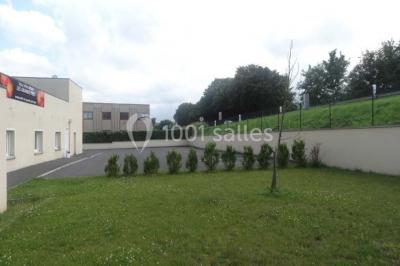 Bâtiment beige à un étage avec des fenêtres blanches, entouré d'une pelouse et d'arbustes, sous un ciel partiellement…