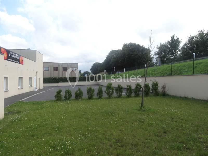 Pelouse avec jeune arbre, haie de buissons, bâtiment beige à gauche et talus verdoyant en arrière-plan.