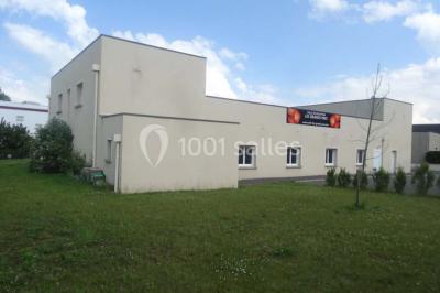 Bâtiment beige à un étage avec des fenêtres blanches, entouré d'une pelouse et d'arbustes, sous un ciel partiellement…