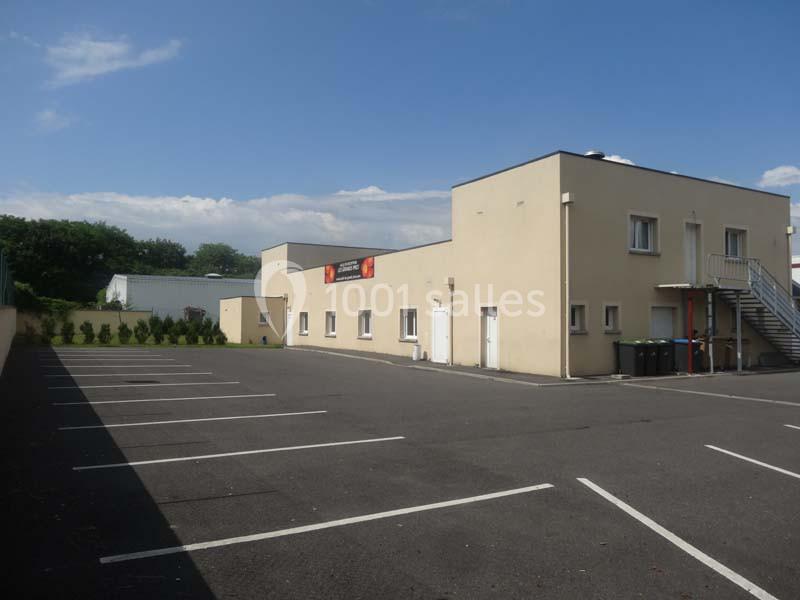 Parking vide devant un bâtiment beige à un étage, avec quelques arbres en arrière-plan sous un ciel dégagé.