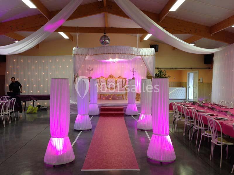 Salle de réception décorée avec un trône doré, tapis rouge, colonnes lumineuses roses et tables dressées.